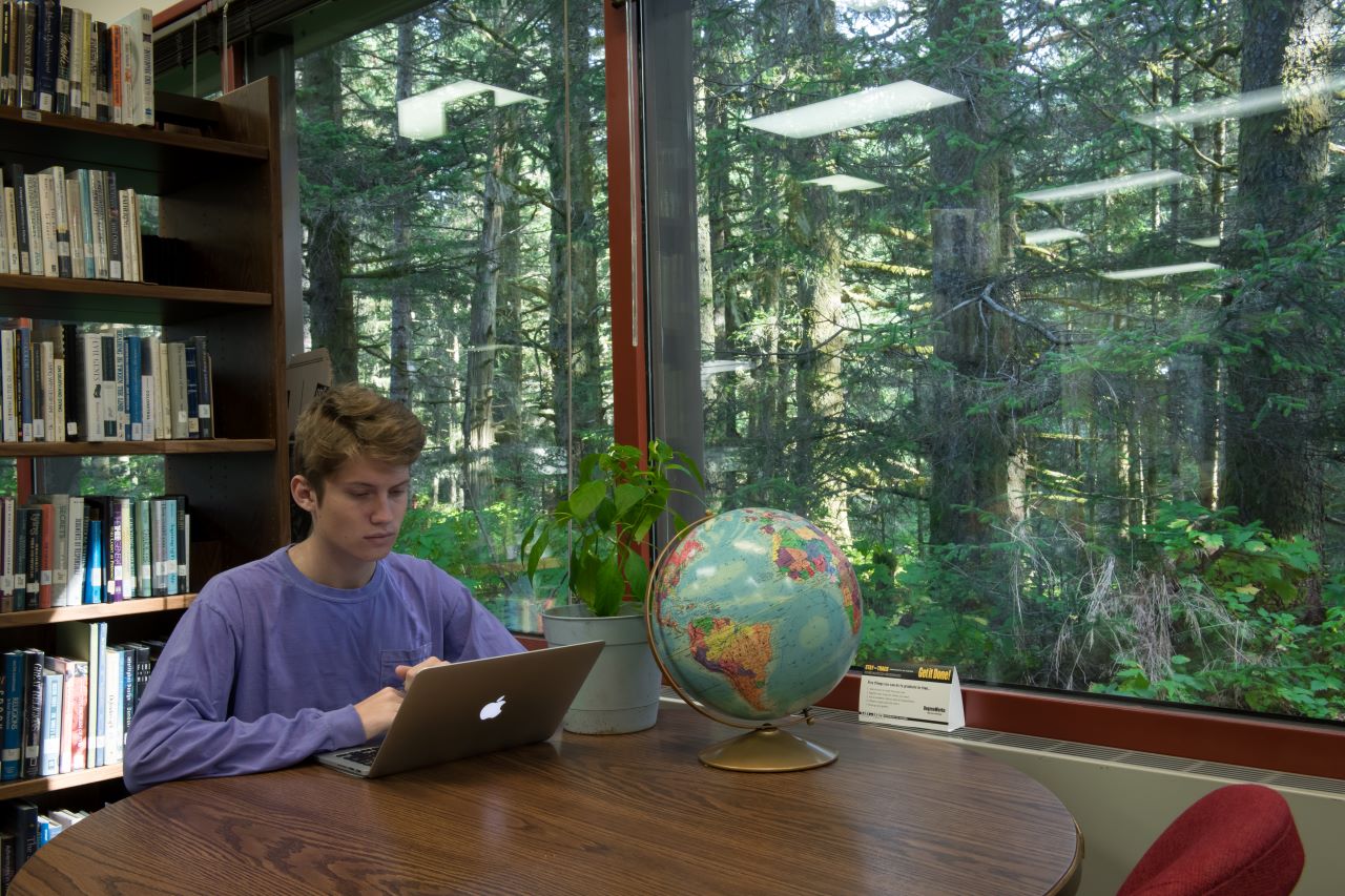 Student working near a window on a laptop on a library table next to a globe