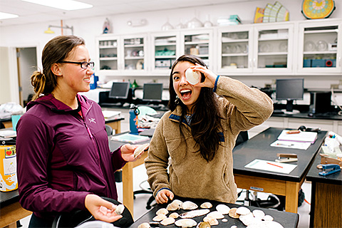 2 students in a science class sorting clam shells with one holding a shell in front of her eye