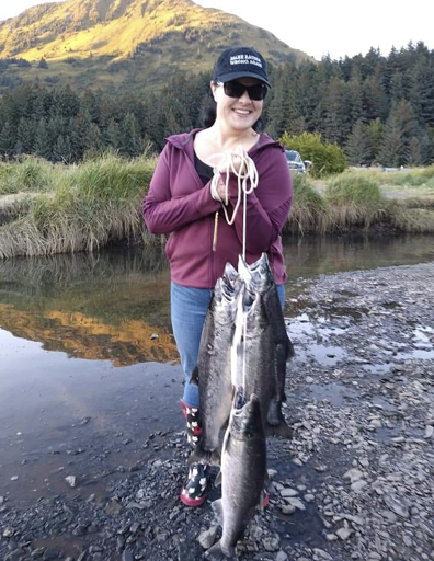 Jacelyn Keys in boots, jeans, and hoodie standing on a river bank holding a string of salmon