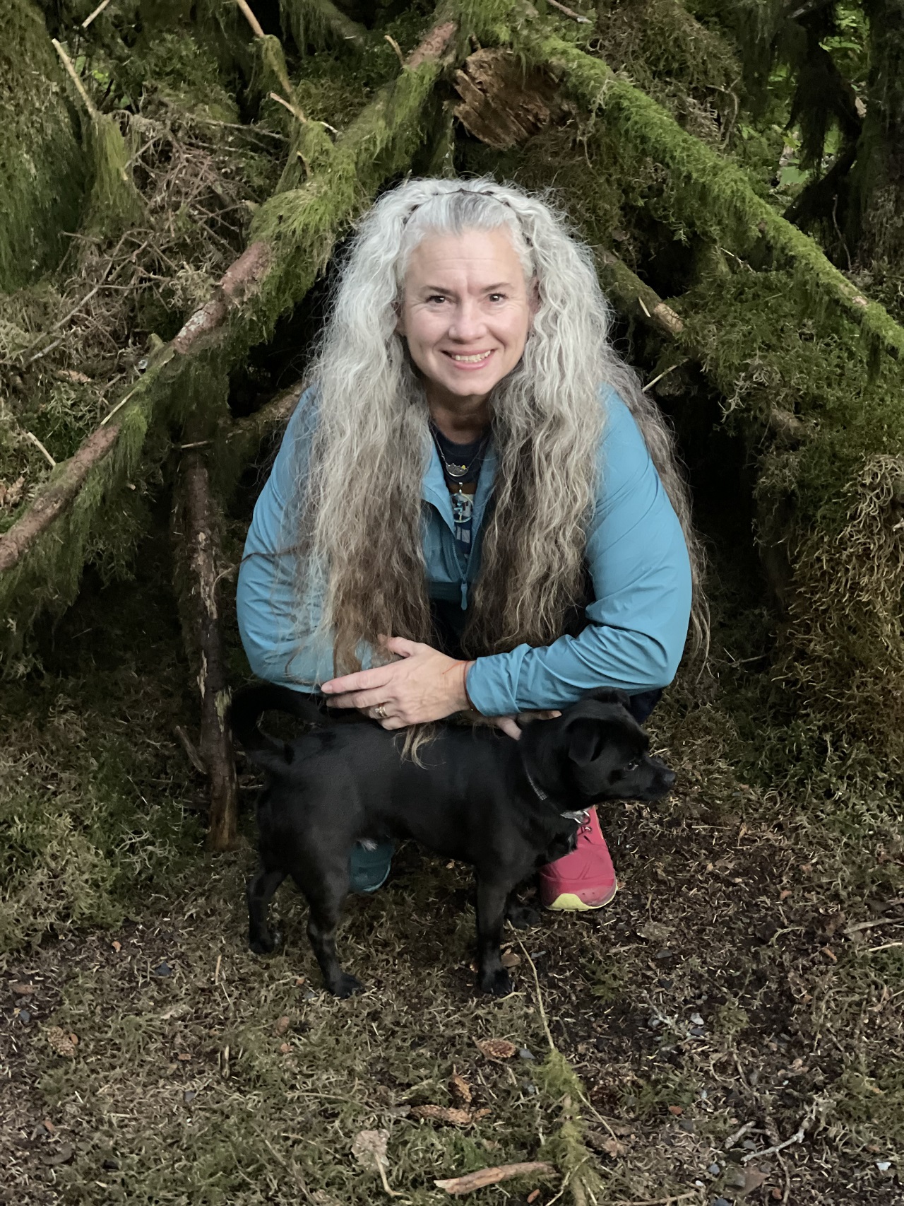 Woman with a small black dog in a forest in front of a makeshift shelter made of mossy limbs