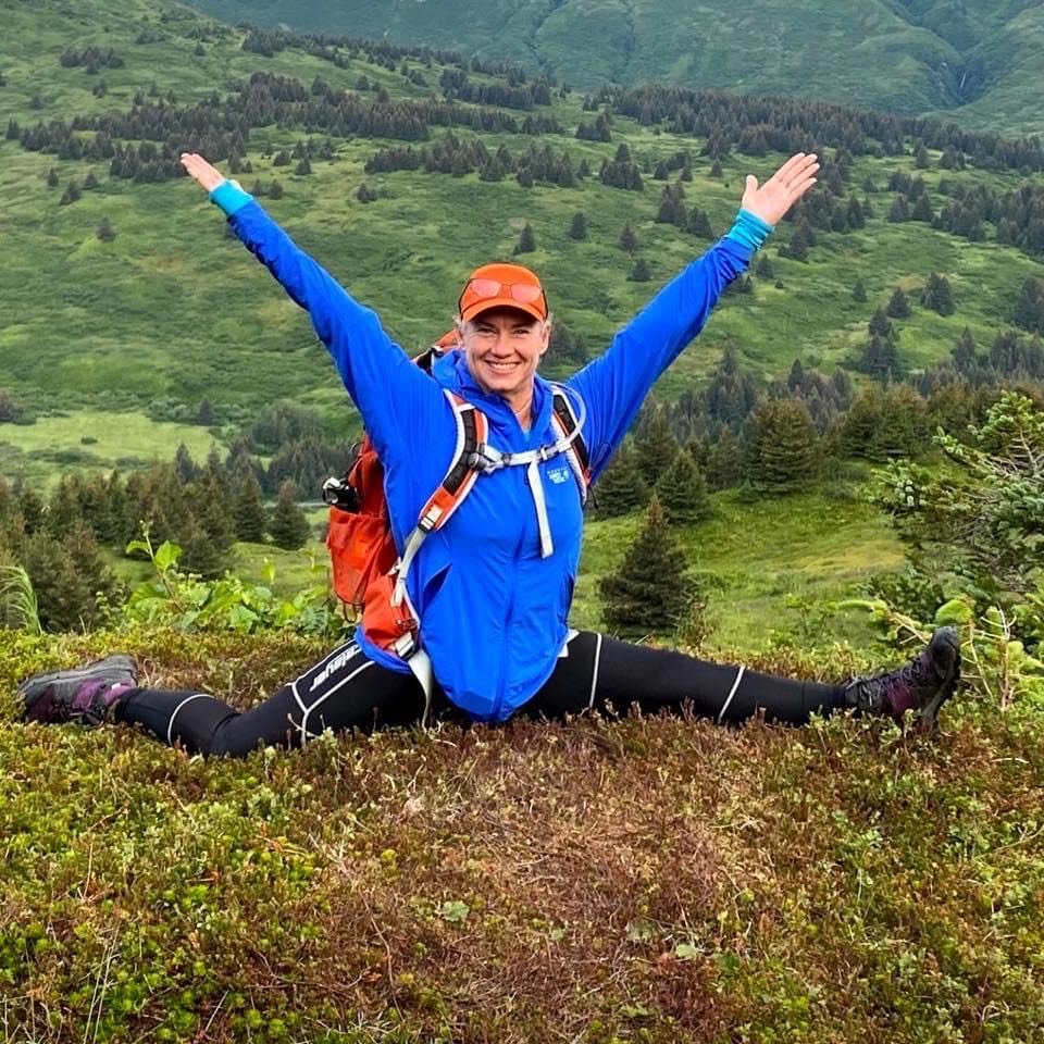 woman wearing a blue jacket and orange ball cap doing a split on top of a mountain