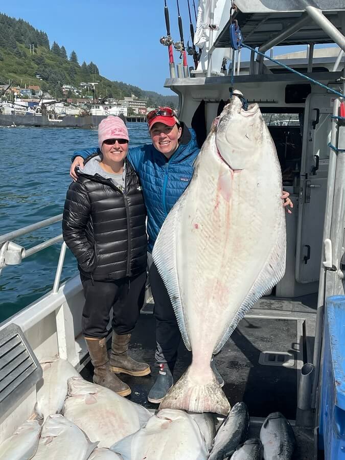 Kim Saunders and Kate Korrow standing on a boat next to a large halibut hanging from rigging with many halibut and salmon on the deck