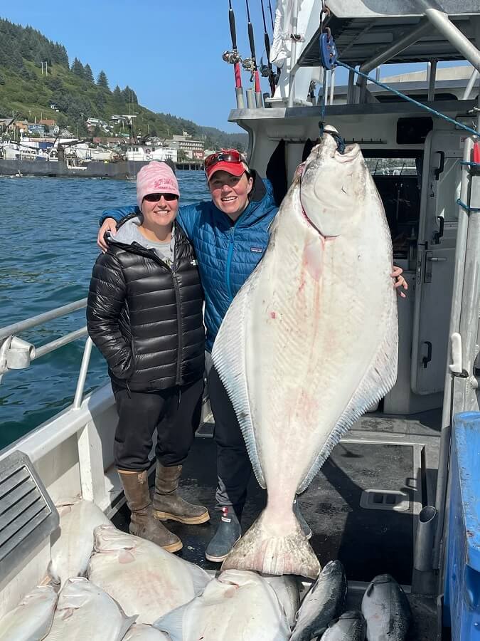 Two women standing on a boat with a large halibut hanging from rigging with other fish on the deck